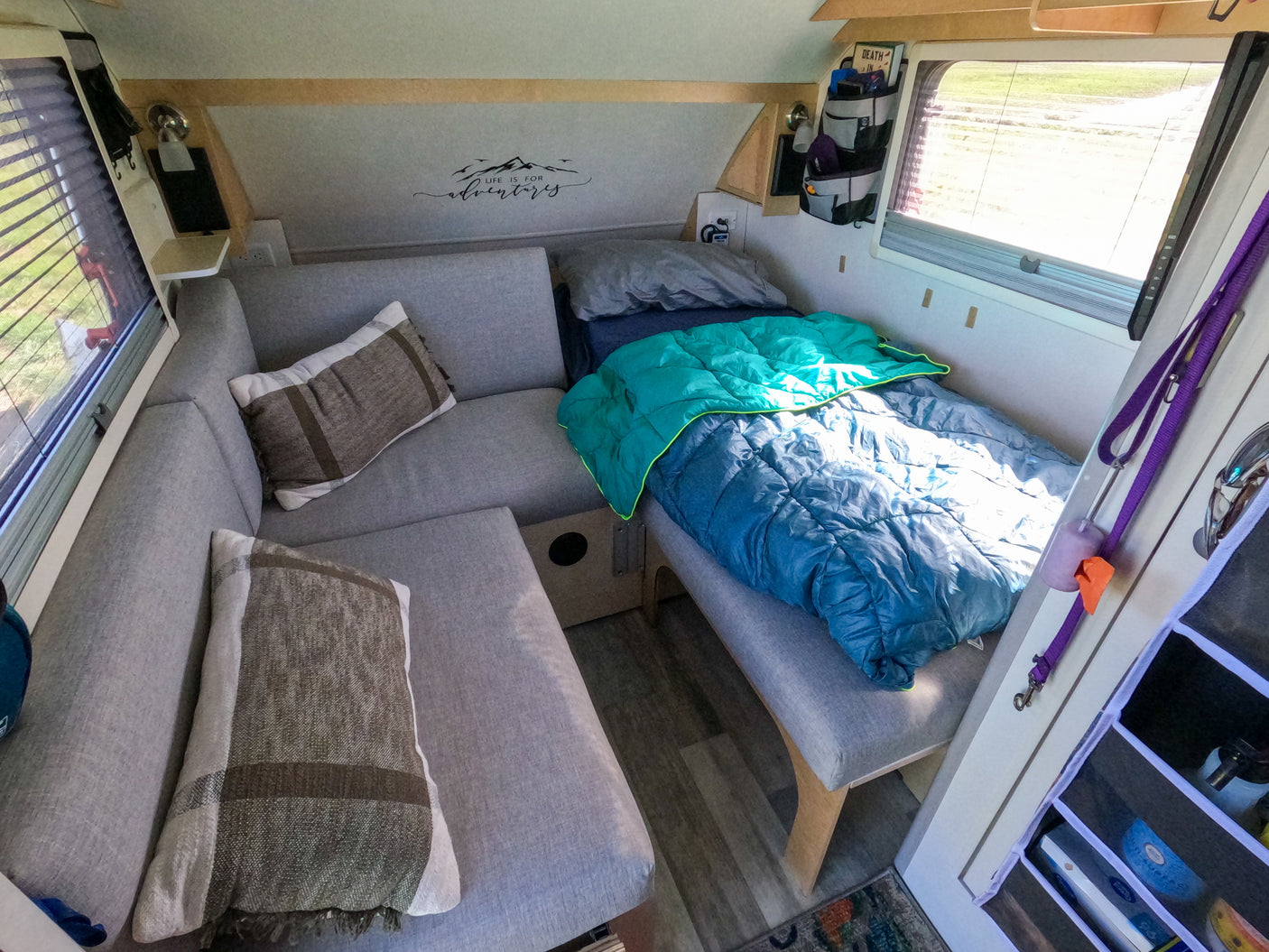This RV rental's interior features comfy gray seating, a bed with blue bedding, and "Life is for adventures" wall art.