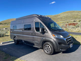 A grey Winnebago Solis RV rental camper van parked on asphalt with hills in the background under a blue sky.