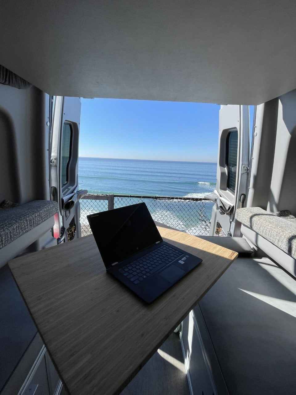 This RV rental features a mobile office setup with a laptop on a table, overlooking a stunning ocean and beach scene from the open rear doors. — photo 7