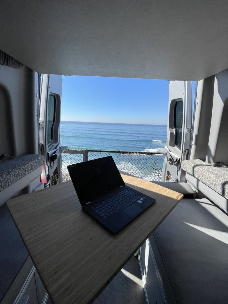 This RV rental features a mobile office setup with a laptop on a table, overlooking a stunning ocean and beach scene from the open rear doors. — photo 7