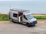Scenic rv rental: a light gray Mercedes Sprinter camper van parked by the ocean, with its side door open showing a wooden interior.