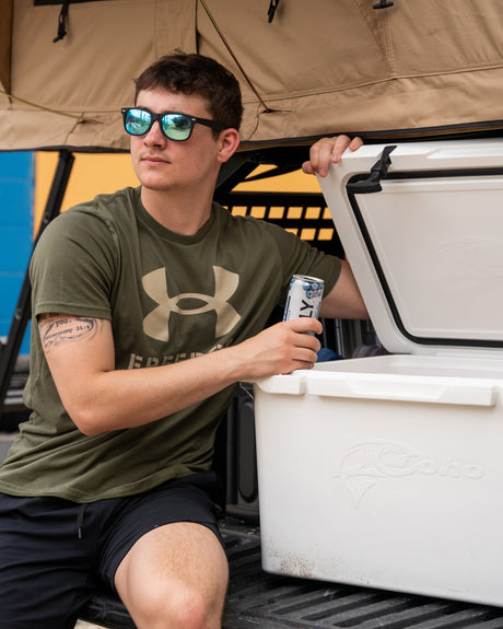 Young man in mirrored shades sips a seltzer from his cooler, relaxing under a rooftop tent during his RV rental.