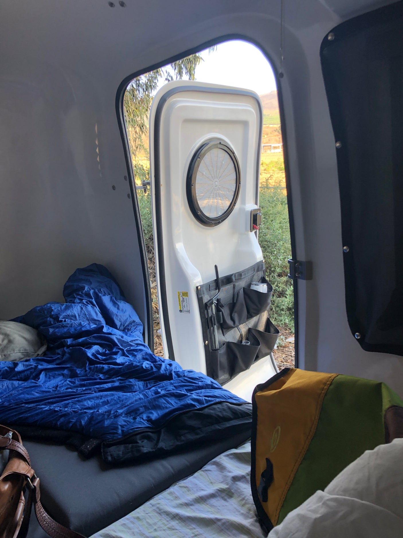 Inside an rv rental camper, a blue sleeping bag on the bed, with the door open to a view of hills and nature.