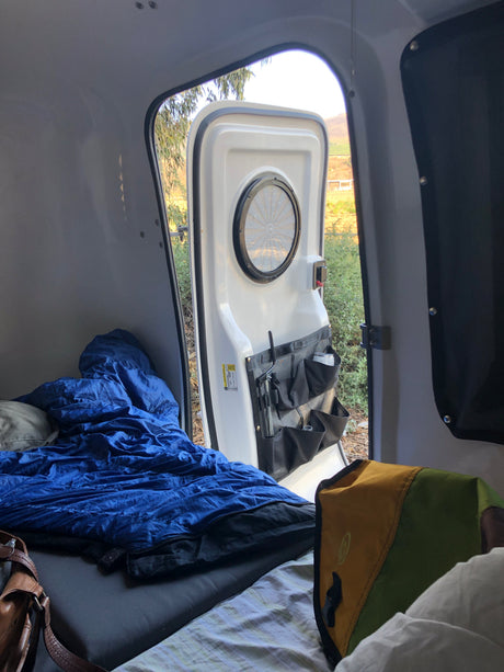 Inside an rv rental camper, a blue sleeping bag on the bed, with the door open to a view of hills and nature.