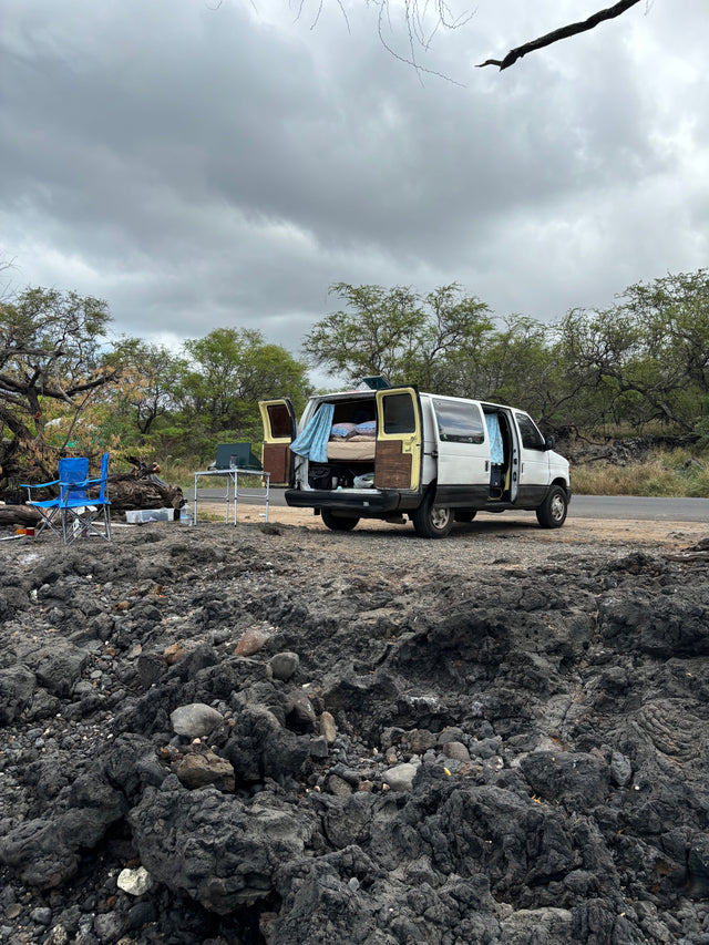 RV rental camper van parked on rugged lava rock, with open doors, bed, and camp setup, under an overcast sky.