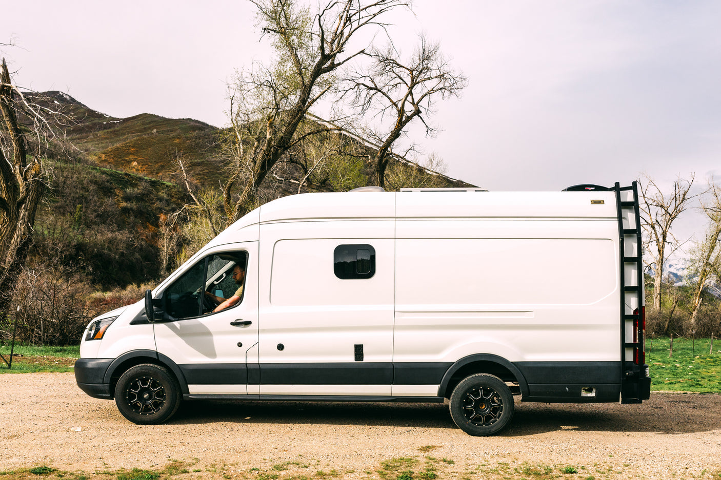 White RV rental van on a dirt road, driver visible. Scenic mountains & trees. Perfect for adventure & van life.