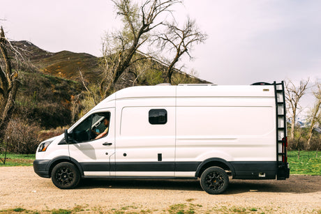White RV rental van on a dirt road, driver visible. Scenic mountains & trees. Perfect for adventure & van life.