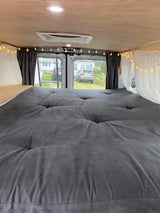 Experience a comfortable RV rental! This custom van interior features a plush grey tufted bed, wood walls, and cozy lights.