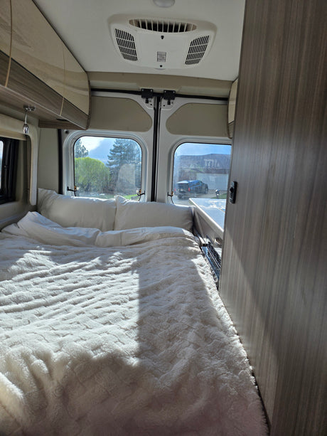 Cozy RV rental interior with a comfy bed, overhead storage, and bright windows offering a view of the outdoors.