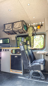 Inside an rv rental: custom camper van interior with a kitchen, refrigerator, flexible seating, and overhead storage. View of forest.