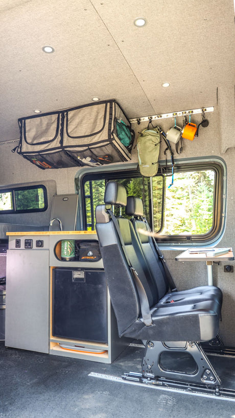 Inside an rv rental: custom camper van interior with a kitchen, refrigerator, flexible seating, and overhead storage. View of forest.
