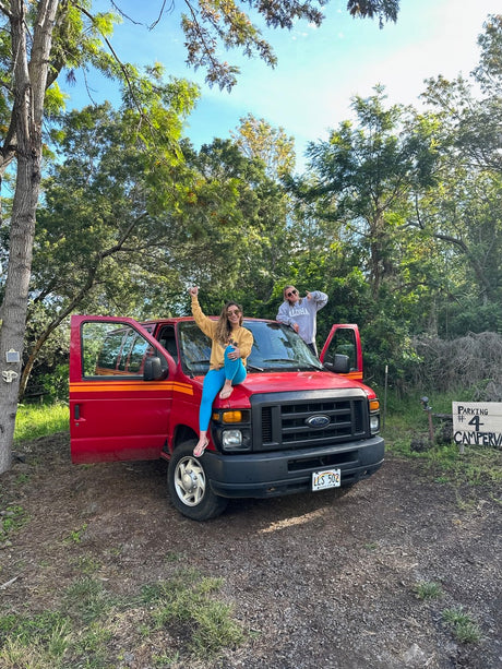 Two joyful women pose with their red Ford RV rental van in nature. One sits on the hood, the other leans by the open door.