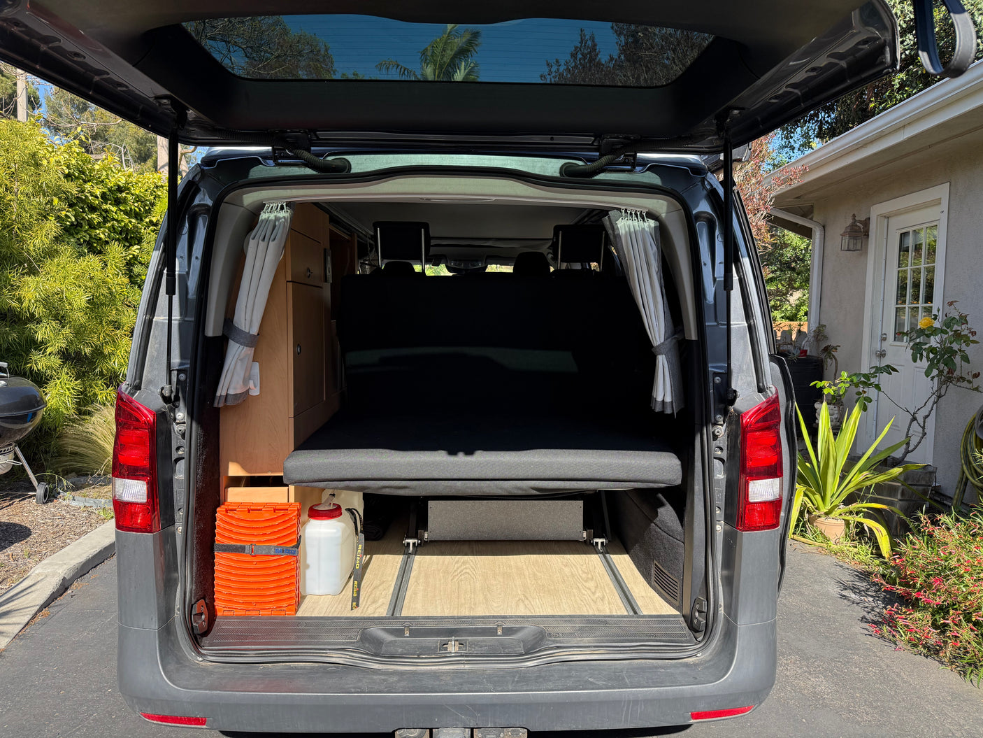 Open tailgate of an RV rental camper van, showing a compact interior with a bed, wood floor, and built-in storage.