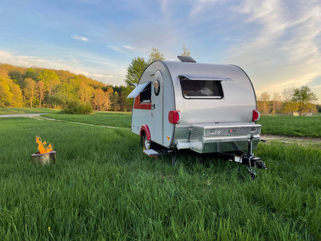 Teardrop RV rental parked in a vibrant green field with a roaring campfire; surrounded by trees under a soft sky.