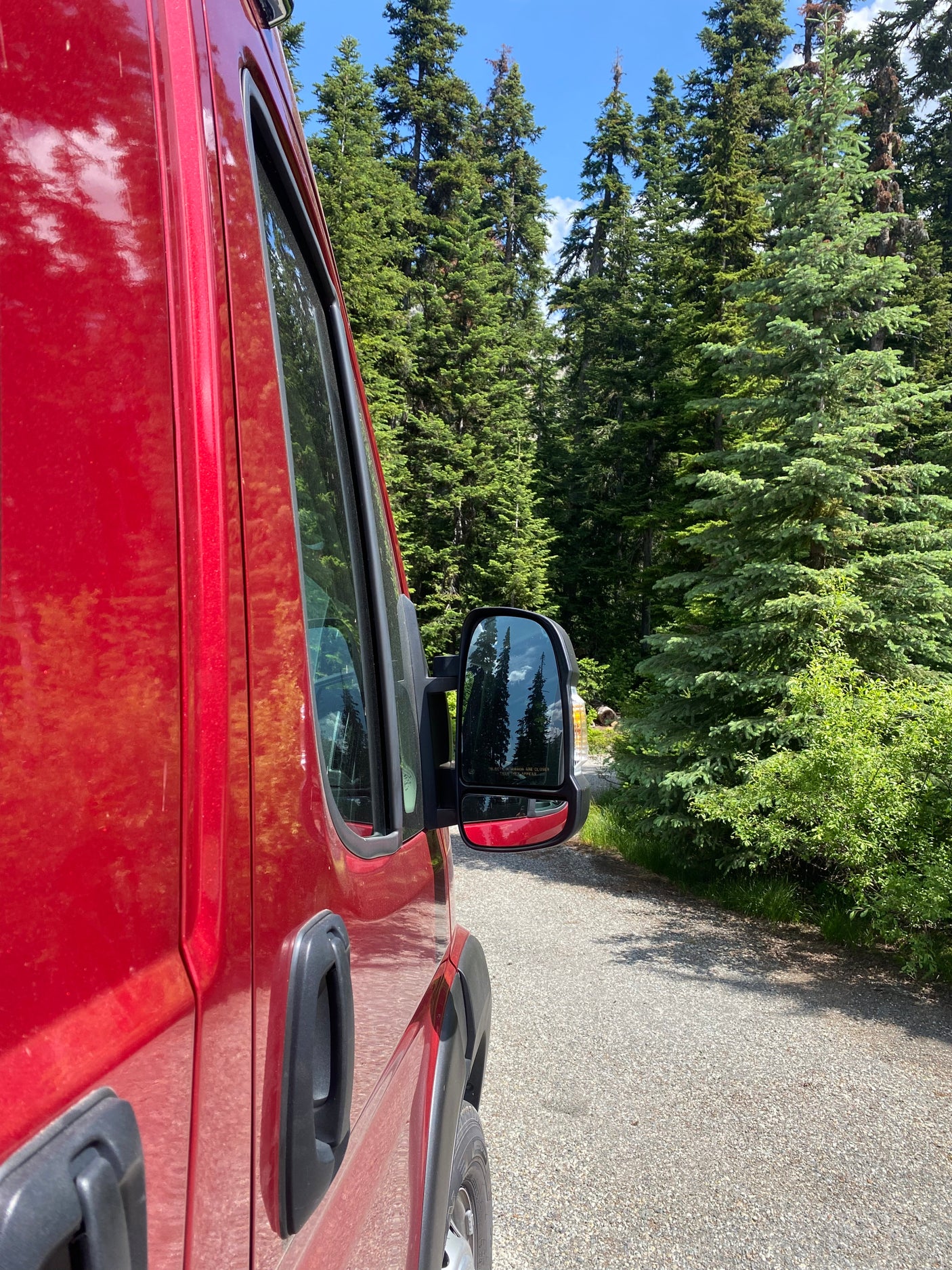 Side view of a red RV rental camper van parked on a gravel forest road, surrounded by tall evergreen trees.