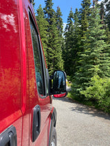 Side view of a red RV rental camper van parked on a gravel forest road, surrounded by tall evergreen trees.