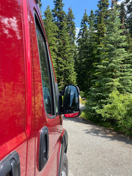 Side view of a red RV rental camper van parked on a gravel forest road, surrounded by tall evergreen trees.