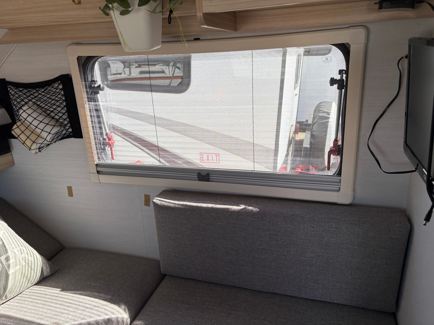 Cozy RV rental interior: bright window with screen, soft gray seating, storage net, and TV. Enjoy comfort on your road trip. — photo 7
