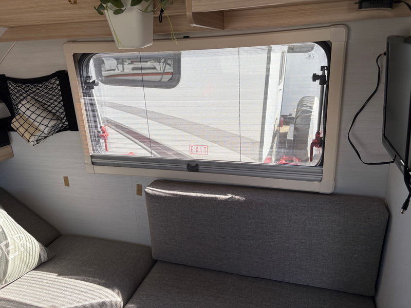 Cozy RV rental interior: bright window with screen, soft gray seating, storage net, and TV. Enjoy comfort on your road trip. — photo 7