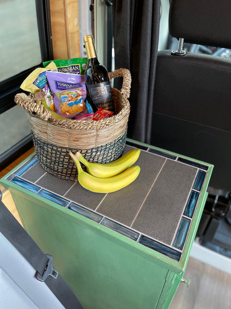 An RV rental welcome basket with Hawaiian snacks, wine, and bananas on a tiled cabinet. Perfect for travel.