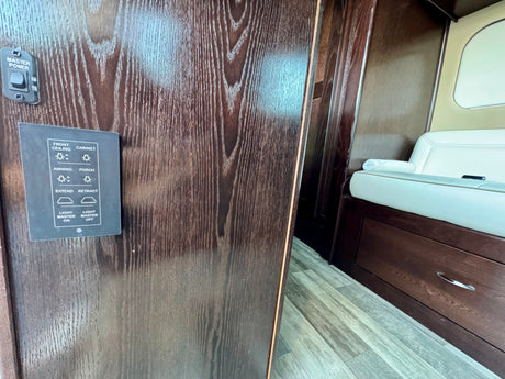 RV rental interior: Dark wood wall with a master power switch and control panel for lighting and awning, beside cream seating.