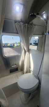 Interior of an RV rental bathroom with a toilet, shower, windows, and bright ceiling light, showing compact design.