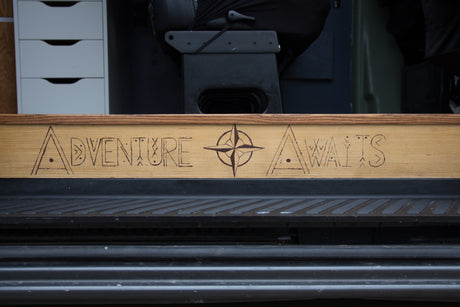 RV rental interior: a custom wood panel with 'ADVENTURE AWAITS' and compass art. Ready for your next travel journey.