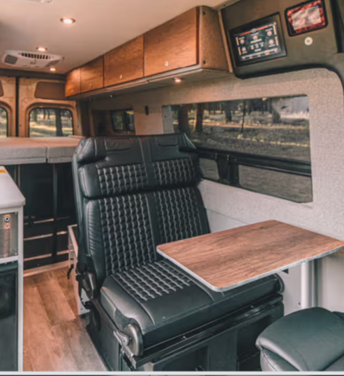 RV rental interior featuring a plush black seat with a wood table, overhead cabinets, and a bed. Ideal for camper van travel.