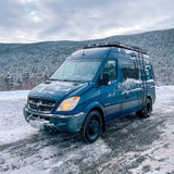 Blue Dodge Sprinter camper van (RV rental) covered in snow, parked on a winter mountain road with snowy trees.