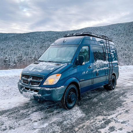 Blue Dodge Sprinter camper van (RV rental) covered in snow, parked on a winter mountain road with snowy trees.