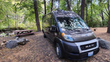 Dark gray camper van as an rv rental parked at a tranquil wooded campsite, with a picnic table & stream nearby.