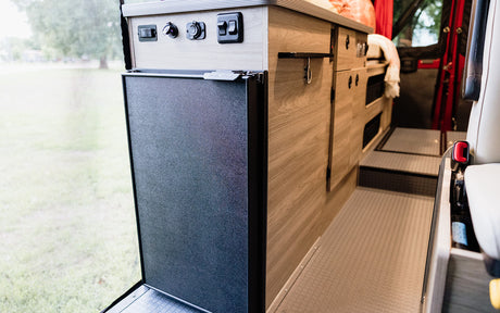 RV rental: Inside a camper van with a Nova Kool refrigerator, control panel, and wood storage cabinets.