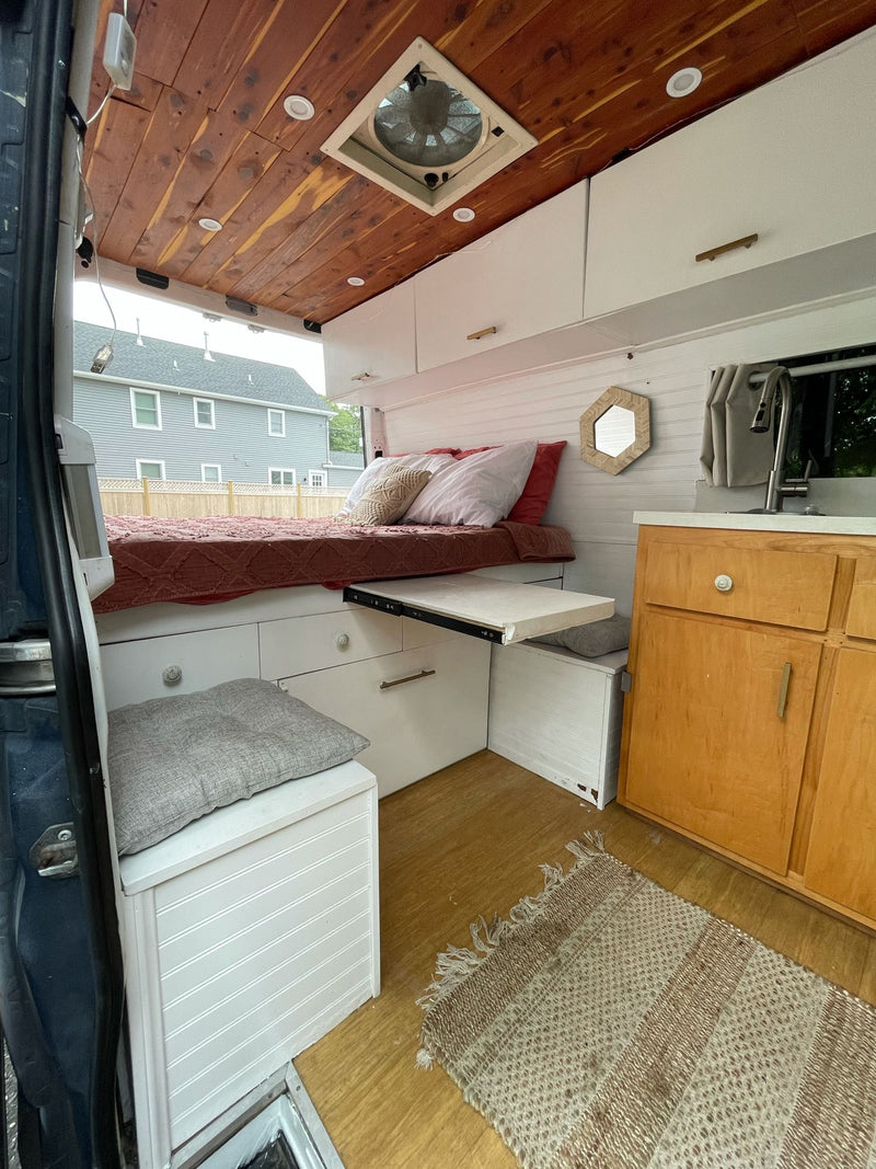 Explore this modern RV rental camper van interior. Features a cozy bed, pull-out table, kitchen, storage, and wood ceiling. — photo 7