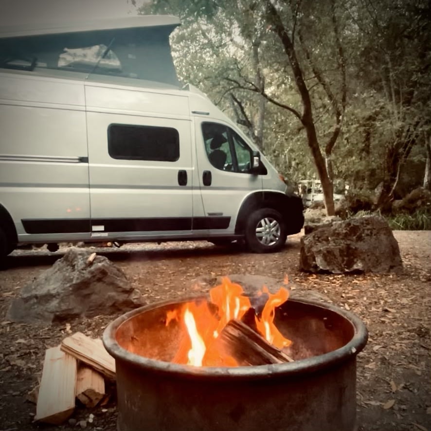 RV rental adventure: a white camper van parked in a serene, treed campsite with a warm campfire burning in the foreground.