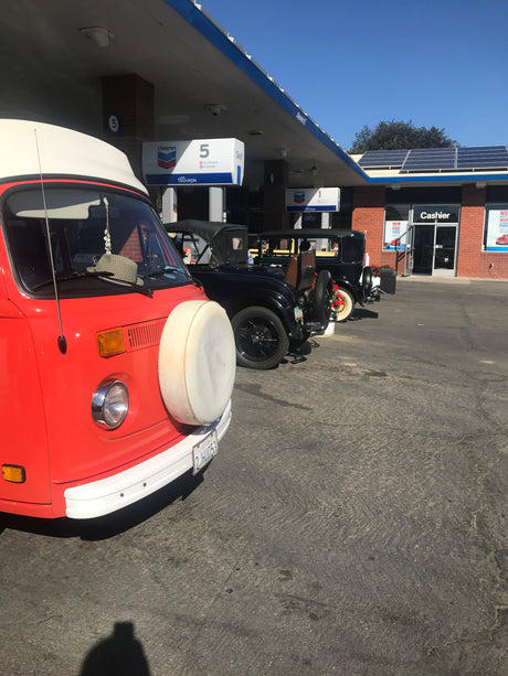 An orange vintage RV (possibly an RV rental) and classic cars are parked at a Chevron gas station with solar panels.