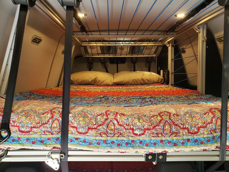 Comfortable bed inside an RV rental, featuring a vibrant patterned comforter, pillows, and custom overhead storage.