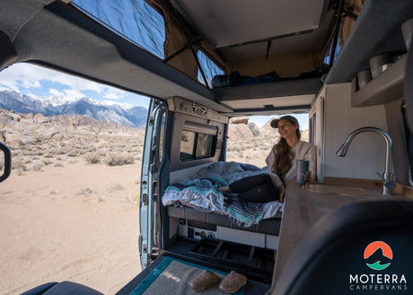  and gear storage for comfortable adventures.|Cozy RV rental bed with open back doors revealing stunning river and mountain views. Your ultimate camper van escape.|This RV rental offers a woman comfy van life with breathtaking mountain views and kitchen access. Escape today!|A sleek gray Moterra Sprinter campervan
