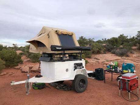 Overland RV rental setup: camping trailer with rooftop tent and outdoor kitchen amidst red desert. Ready for adventure!