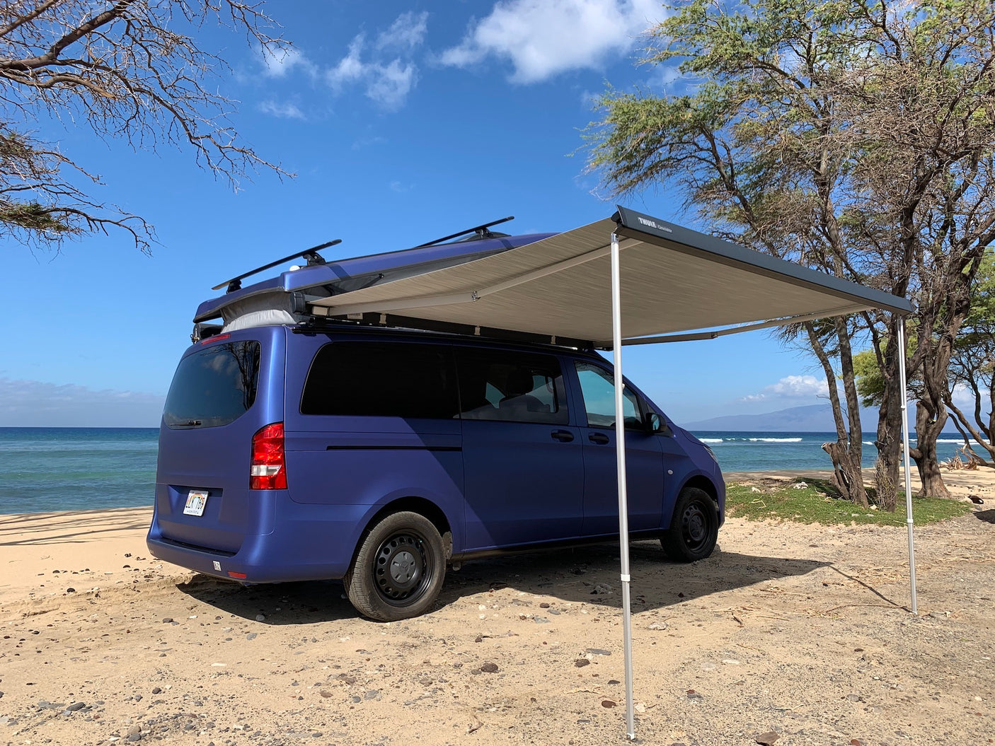 Blue rv rental camper van with awning parked on a sandy Hawaii beach by the ocean under blue skies.