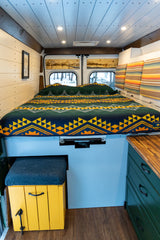 Cozy camper van RV rental interior: bed with patterned blanket, wood paneling, and storage.