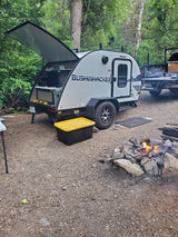 A Bushwhacker teardrop **rv rental** at a forest campsite. Its rear galley is open, with a truck and campfire nearby.
