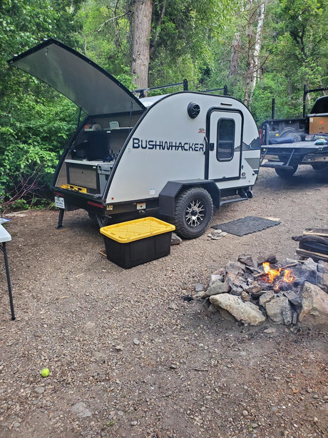 A Bushwhacker teardrop **rv rental** at a forest campsite. Its rear galley is open, with a truck and campfire nearby.