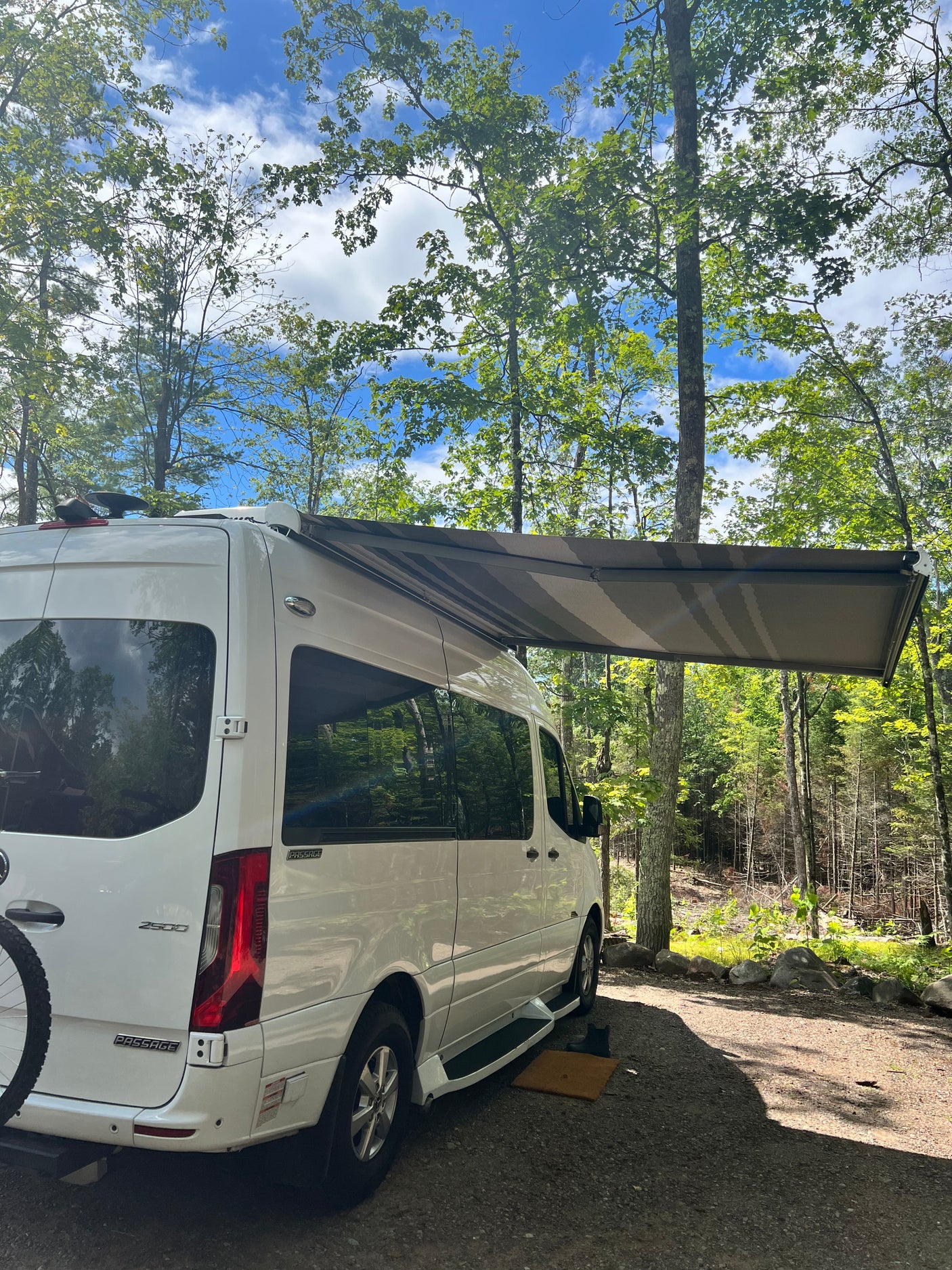 White RV rental with awning extended, parked in a sunny wooded campsite. Perfect for a relaxing nature travel escape.