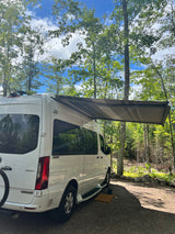 White RV rental with awning extended, parked in a sunny wooded campsite. Perfect for a relaxing nature travel escape.