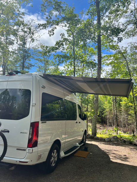 White RV rental with awning extended, parked in a sunny wooded campsite. Perfect for a relaxing nature travel escape.