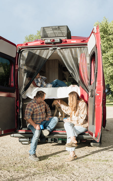 Family enjoying their **RV rental**, with adults and dog at the open back of a red van, and a child on the bed inside.