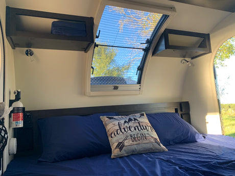 Cozy RV rental interior shows a blue bed, adventure pillow, overhead storage, & a bright skylight with a screen & outdoor view.