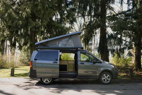 Gray pop-top camper van rv rental in a lush park setting. Side door open, showcasing a cozy, custom-built wooden interior.
