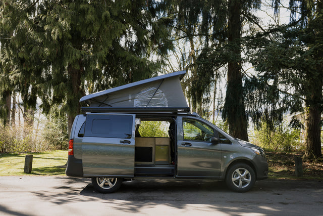 Gray pop-top camper van rv rental in a lush park setting. Side door open, showcasing a cozy, custom-built wooden interior.