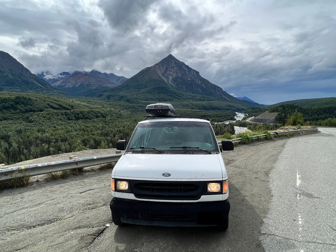 Scenic view with an rv rental: a white Ford van parked roadside against a backdrop of mountains, forests, and a river.